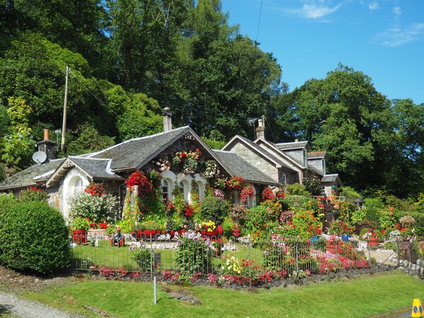 Ein schönes Steinhaus, umgeben von einem üppigen Garten voller bunter Blumen, mit einem reich verzierten Blumenbeet und grünen Bäumen im Hintergrund, an einem sonnigen Tag.